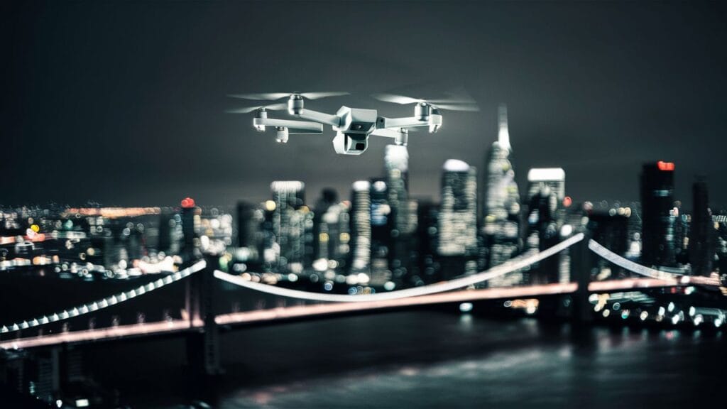 A drone flying at night, illuminated by its own lights, against a dark sky with city lights in the background. The whole scene answers the question that can drones fly at night?